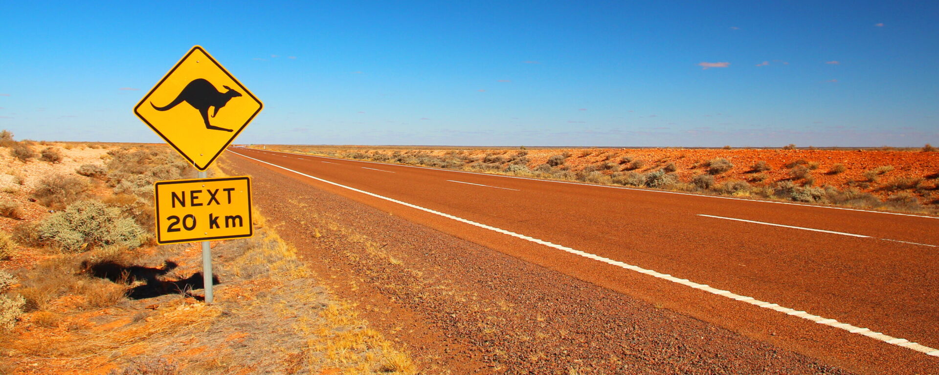 Ikonisches gelbes Warndreieck-Schild mit springendem Känguru und "NEXT 20 km" auf roter Outback-Straße in Australien – Känguru-Gefahr im australischen Inland. 