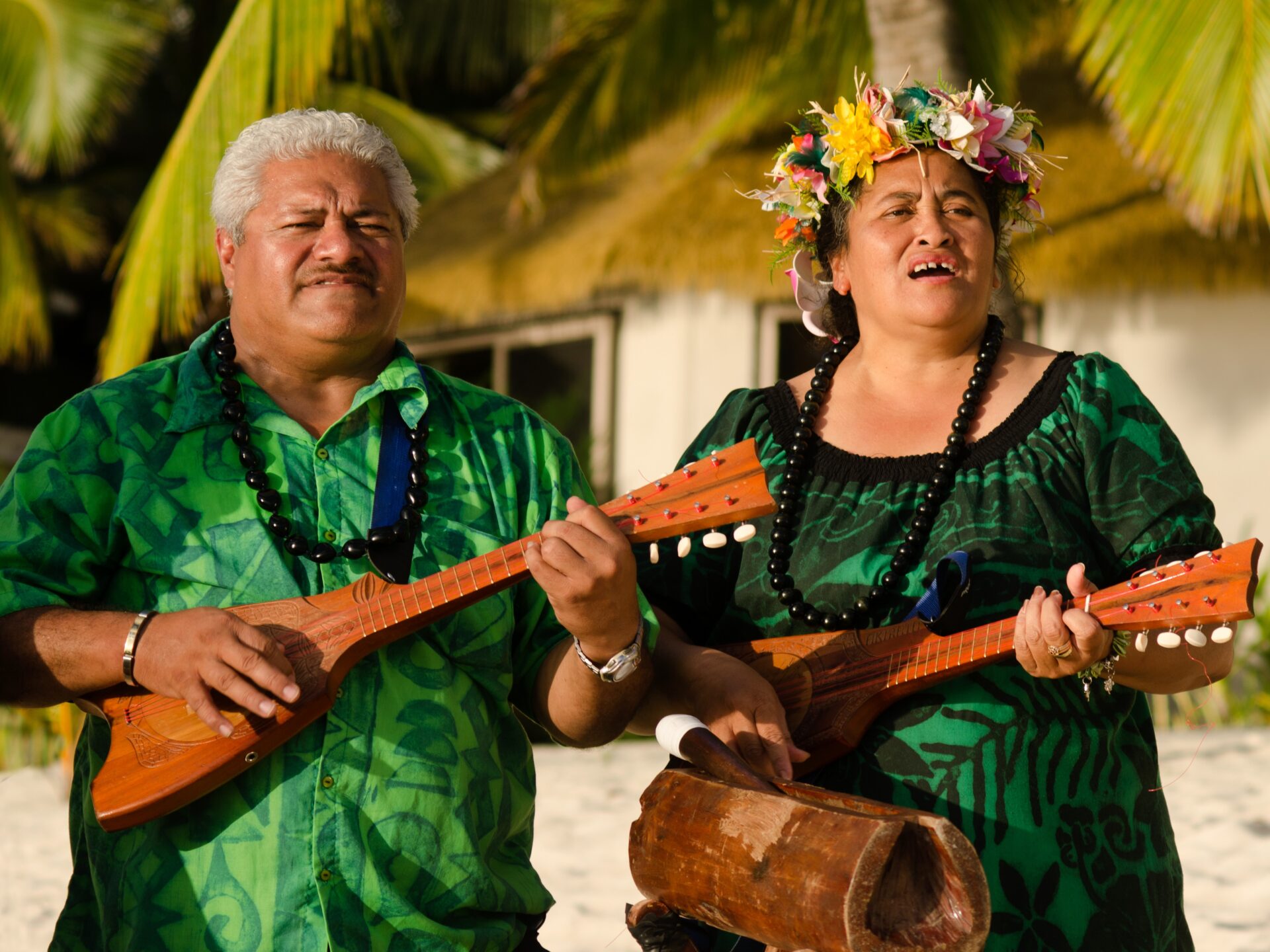 Lächelndes polynesisches Paar in traditioneller Kleidung mit Blumenkränzen spielt Ukulele vor traditionellem Haus auf den Cookinseln – authentische Begrüßungsmusik und Kultur. 