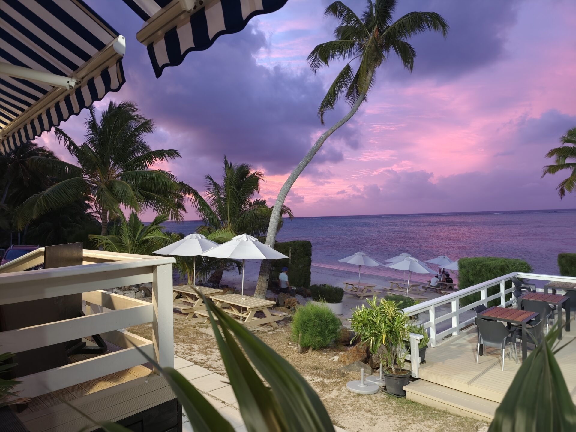 Beeindruckender Sonnenuntergang an der Westküste von Rarotonga mit rosa-purpur Himmel, Palmen, weißem Sandstrand, Sonnenschirm und Bungalow.
